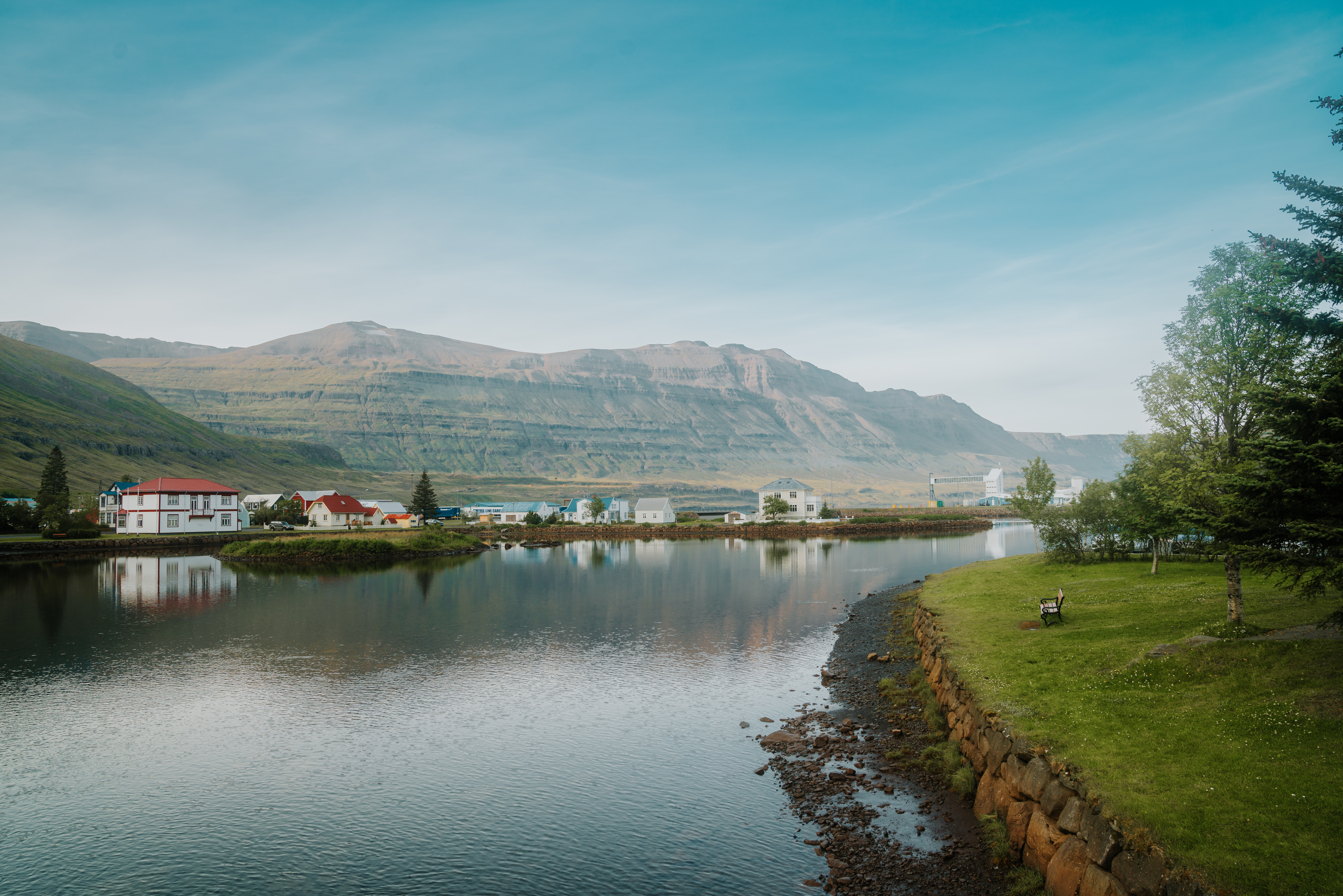 A calm lagoon reflecting white houses with red roofs, backed by layered brown mountains, with a grass bank, trees and a wooden bench on the right